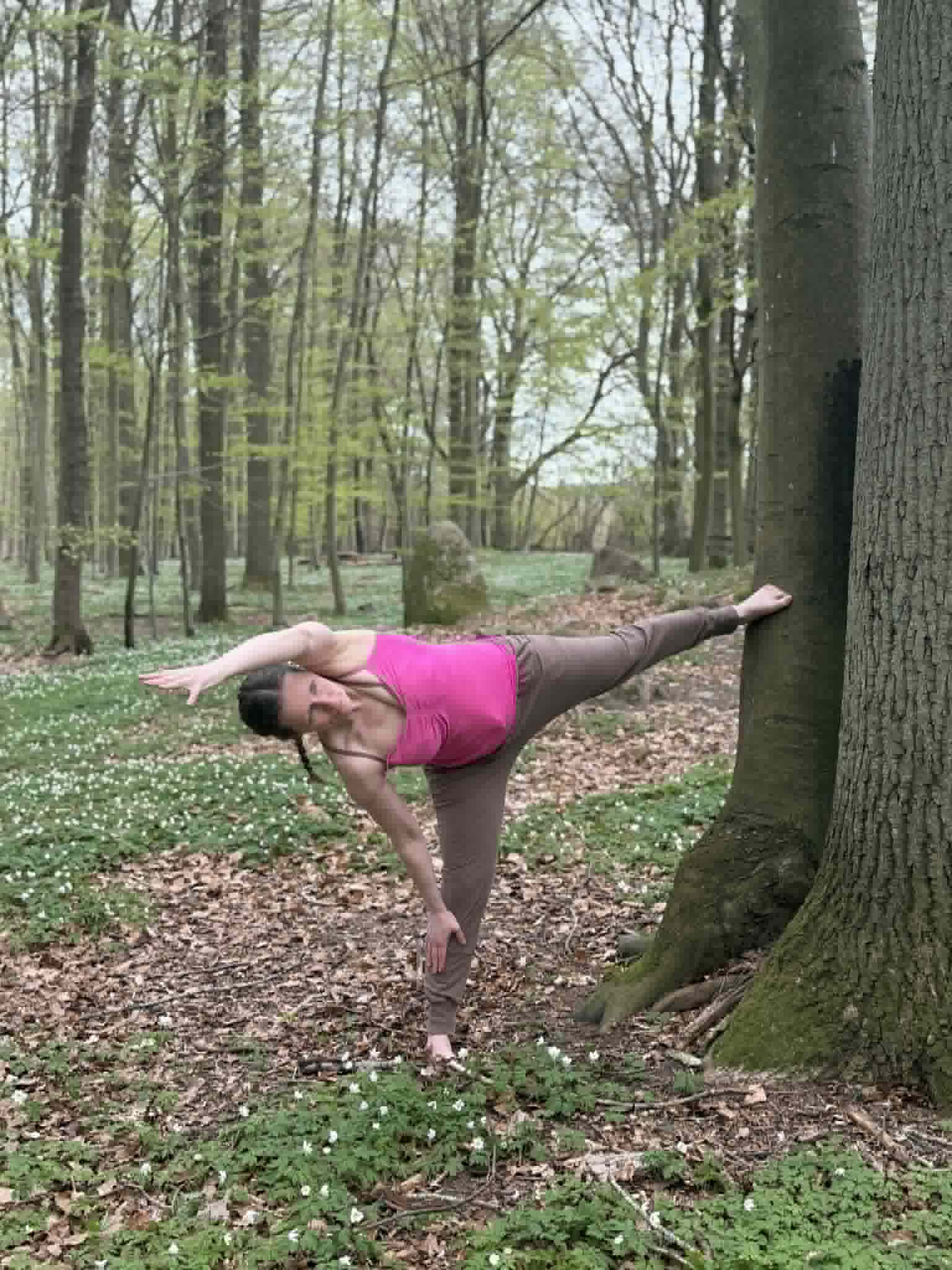 Susan laver yoga med benet strakt op ad et træ i skoven
